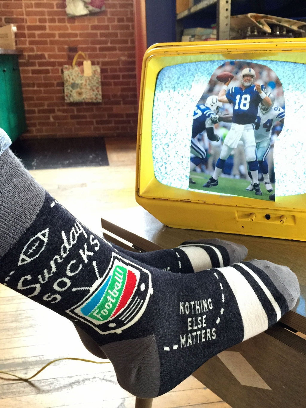 Wearing Blue Q’s Sunday M-Crew Socks featuring football graphics and the phrase "Nothing else matters," a person lounges in sweatpants with warm feet up, watching football on a small yellow TV—making these socks the perfect sports fan gift.