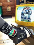 Wearing Blue Q’s Sunday M-Crew Socks featuring football graphics and the phrase "Nothing else matters," a person lounges in sweatpants with warm feet up, watching football on a small yellow TV—making these socks the perfect sports fan gift.