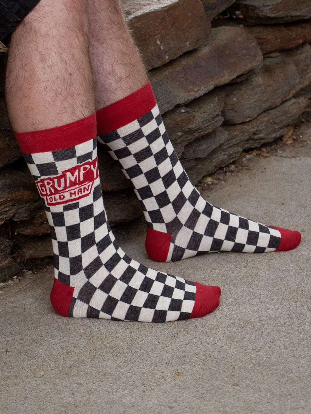 A person wearing Blue Q’s Grumpy Old Man M-Crew Socks—black and white checkered men's novelty socks with red toes, heels, and cuffs—sits outdoors near a stone wall.