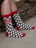 A person wearing Blue Q’s Grumpy Old Man M-Crew Socks—black and white checkered men's novelty socks with red toes, heels, and cuffs—sits outdoors near a stone wall.