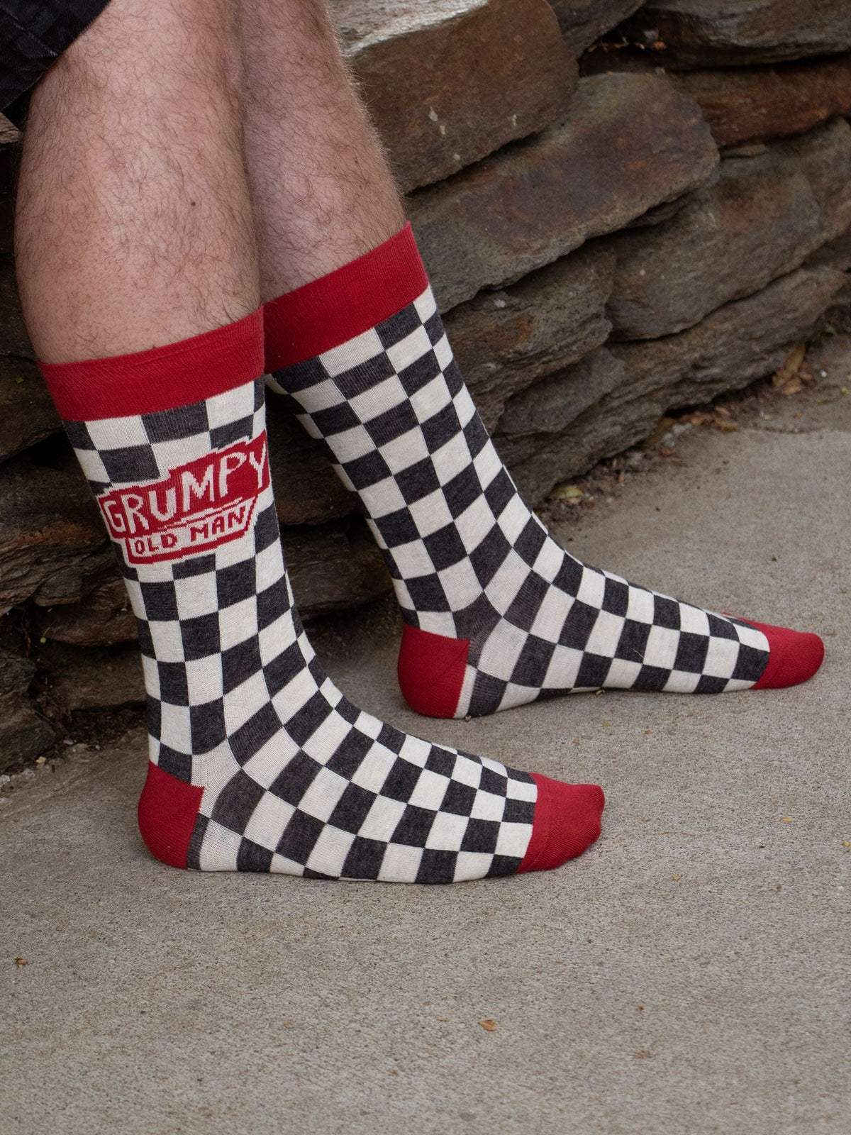 A person wearing Blue Q’s Grumpy Old Man M-Crew Socks—black and white checkered men's novelty socks with red toes, heels, and cuffs—sits outdoors near a stone wall.