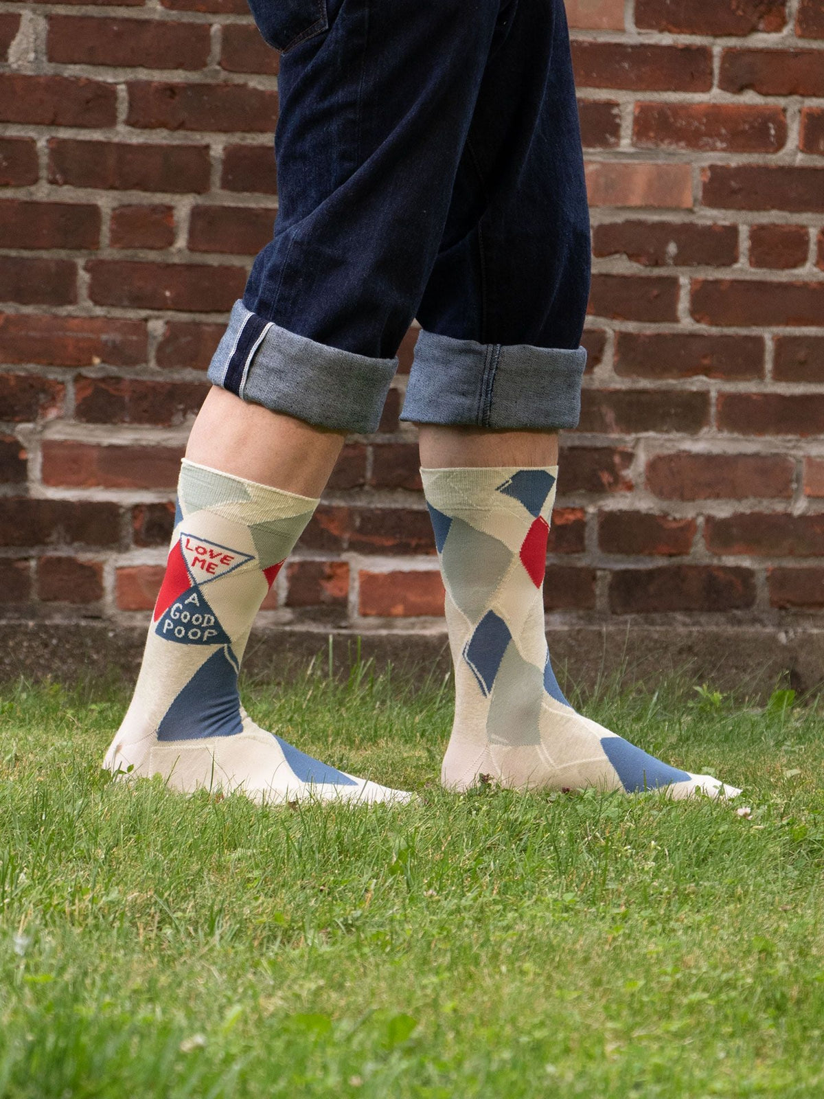 A person stands on grass in rolled-up jeans wearing Blue Q’s Love Me A Good Poop M-Crew Socks, crafted from soft combed cotton in cream with blue and red accents and a woven patch. A brick wall is visible in the background.
