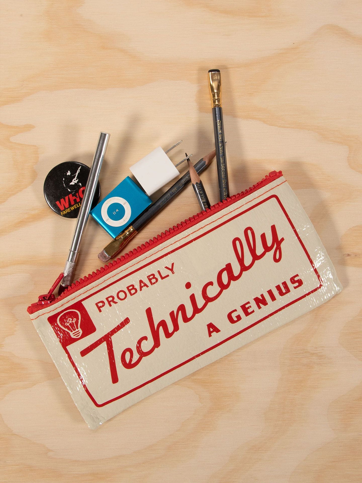 The Blue Q “Probably Technically A Genius” Pencil Case sits open on a wooden surface, surrounded by pens, pencils, a paintbrush, eraser, USB charger, and round pin—essentials for any sharp mind.