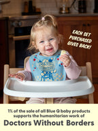 A smiling toddler in a highchair wears a Blue Q “Sorry I’m Crabby. I Slept Like Garbage./Lunch Chucker” terry cotton bib and holds food. Each set purchased supports Doctors Without Borders.
