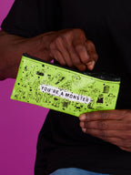 Someone holds a bright green Blue Q "You're A Monster Pencil Case" with cartoon monster doodles and text, set against a purple background.