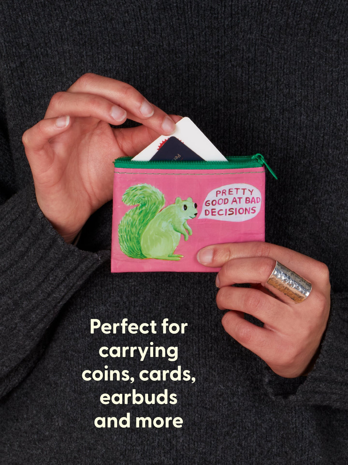 A person holds the Pretty Good At Bad Decisions Coin Purse by Blue Q—a pink pouch featuring a green squirrel and the phrase “Pretty Good at Bad Decisions.” Cards are tucked inside. Text reads: Perfect for carrying coins, cards, earbuds, and more—this quirky Blue Q coin purse is a fun reminder that personal development often begins with some motherly advice (and a great sense of humor).