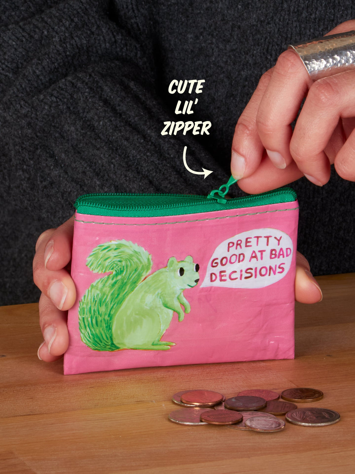 A hand unzips the Pretty Good At Bad Decisions Coin Purse by Blue Q—a small pink pouch featuring a green squirrel and the phrase “Pretty Good at Bad Decisions” for a playful nod to personal growth. Coins are scattered on the wooden table, while an arrow labeled “Cute lil zipper” points to its zipper.