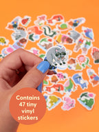 A hand holds a naughty raccoon sticker from the Woodland Critters Caught Smokin’ Playtime Sticker Set, featuring an assortment of cute critters on an orange background with text reading: Contains 47 tiny vinyl stickers.