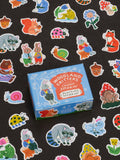 A blue box labeled “Woodland Critters Caught Smokin’ Playtime Sticker Set” by Blue Q is surrounded by colorful stickers featuring cute critters like rabbits, raccoons, foxes, and snails on a black background. Suitable for ages 13 and up.