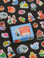 A blue box labeled “Woodland Critters Caught Smokin’ Playtime Sticker Set” by Blue Q is surrounded by colorful stickers featuring cute critters like rabbits, raccoons, foxes, and snails on a black background. Suitable for ages 13 and up.