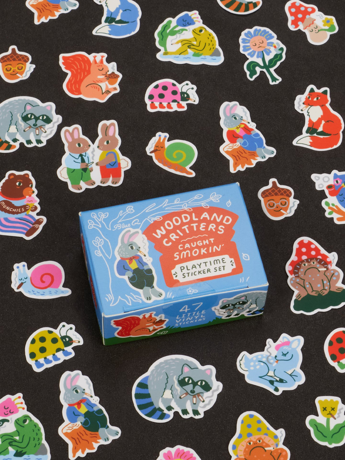A blue box labeled “Woodland Critters Caught Smokin’ Playtime Sticker Set” by Blue Q is surrounded by colorful stickers featuring cute critters like rabbits, raccoons, foxes, and snails on a black background. Suitable for ages 13 and up.