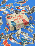 A small cardboard box labeled I’m Really Into Birds Playtime Sticker Set by Blue Q surrounded by numerous bird-shaped stickers on a blue surface.