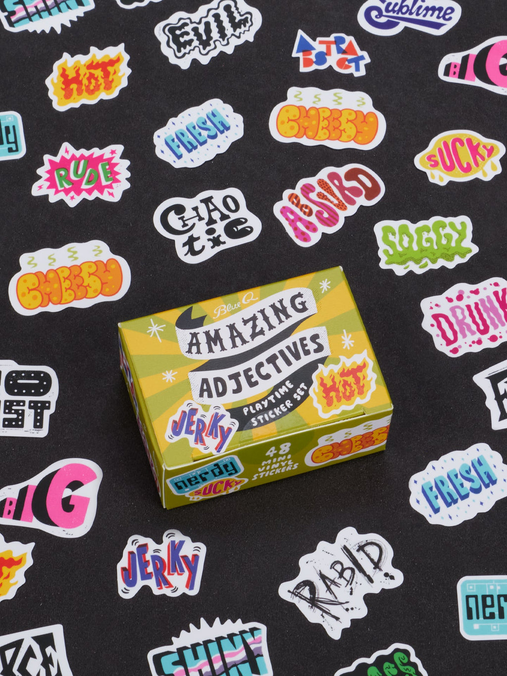 A yellow box labeled Amazing Adjectives Playtime Sticker Set from Blue Q is surrounded by a vibrant array of adjective stickers featuring descriptive words like jerky, hot, rude, soggy, and more—all designed to help modify nouns—set against a black background.
