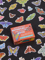 A box of the Blue Q Some Of These Are Butterflies And Some Are Actually Moths Playtime Sticker Set rests on a black surface adorned with vibrant butterfly and moth stickers, making it an ideal pick for lepidopteran enthusiasts ages 13 and up.