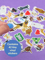 A hand holds a German Shepherd sticker above a pile of Look At These Really Excited Dogs Playtime Sticker Set items on a purple background, with a blue circle overlay stating this set contains 42 tiny vinyl stickers.