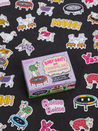 A box labeled “Baby Goats On Trampolines Wearing Sweaters Playtime Sticker Set” by Blue Q rests on a black surface, surrounded by colorful stickers featuring goats on trampolines, geese, playful sound effect words, and more—a perfect novelty gift for ages 13 and up.
