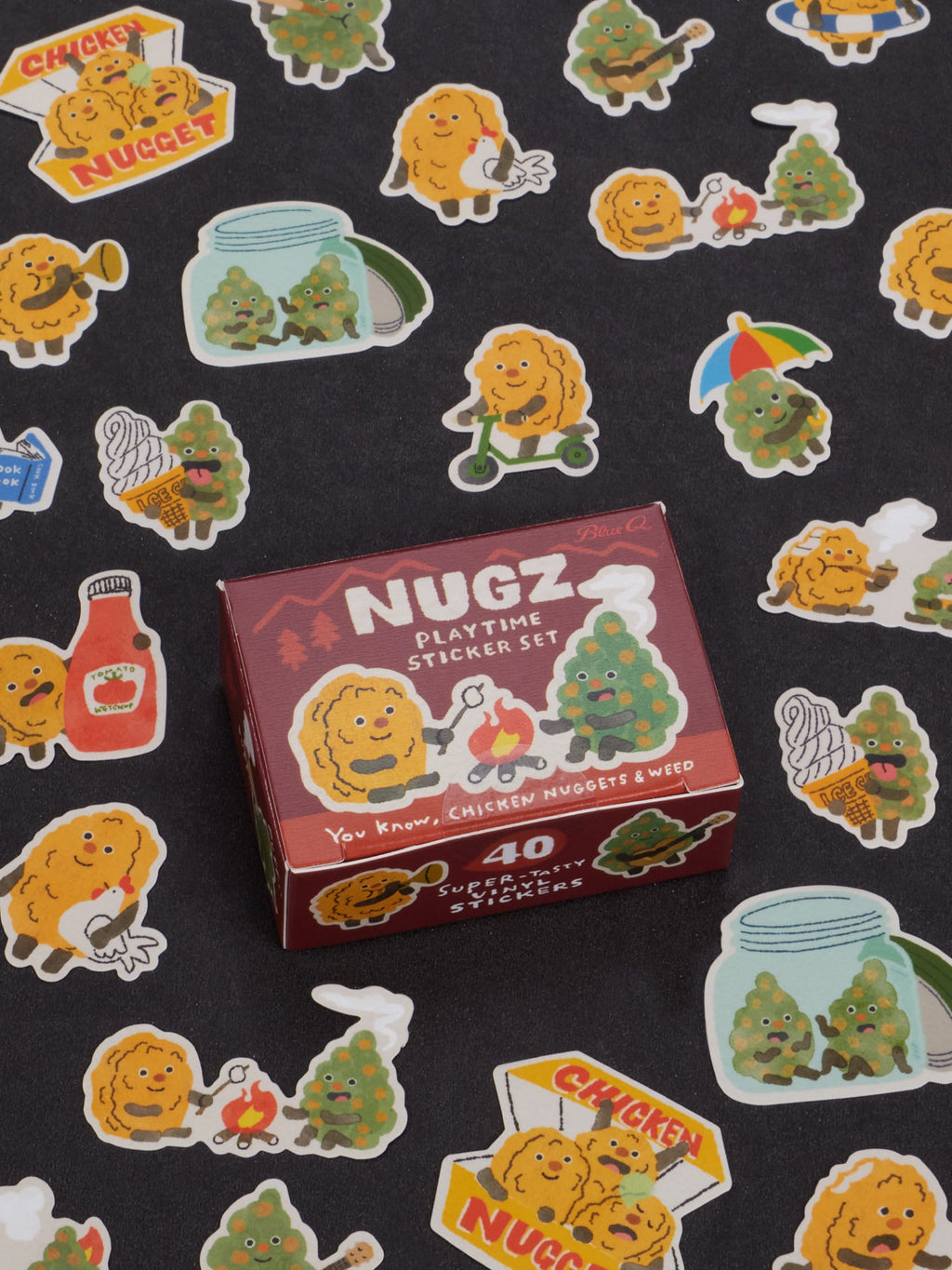 A box labeled Nugz Playtime Sticker Set by Blue Q rests on a black surface scattered with vibrant stickers featuring chicken nuggets, cannabis buds, ketchup, and whimsical cartoon scenes—ideal accessories for teens who want to add a playful touch to their dabber and dipper collections.