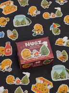 A box labeled Nugz Playtime Sticker Set by Blue Q rests on a black surface scattered with vibrant stickers featuring chicken nuggets, cannabis buds, ketchup, and whimsical cartoon scenes—ideal accessories for teens who want to add a playful touch to their dabber and dipper collections.
