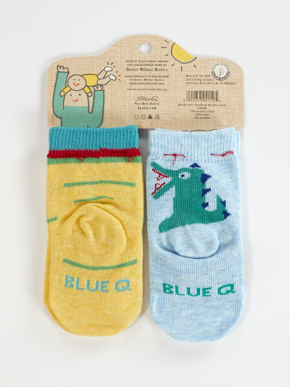 Blue Q's "I Love Your Face/If You Drop Me I'll Remember" baby socks: one yellow with green and red stripes, one light blue with a cartoon dinosaur, made from OCS certified organic cotton in packaging featuring a smiling illustrated character waving.
