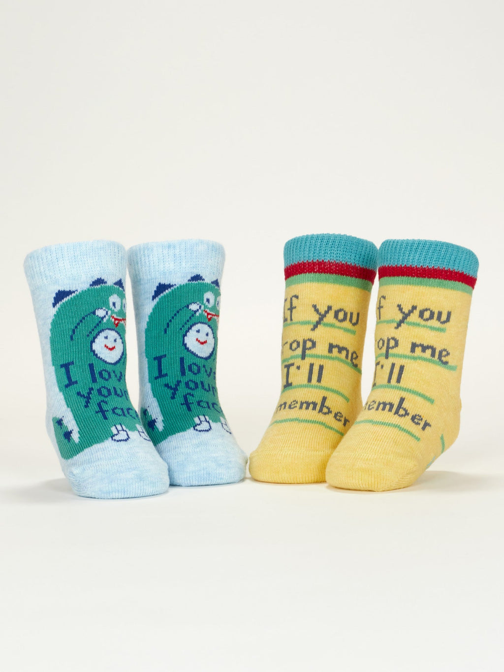 Two pairs of Blue Q's “I Love Your Face/If You Drop Me I'll Remember” baby socks are shown upright: one blue pair with a cartoon bear and message, the other yellow with green trim and a playful phrase. Both are organic cotton.
