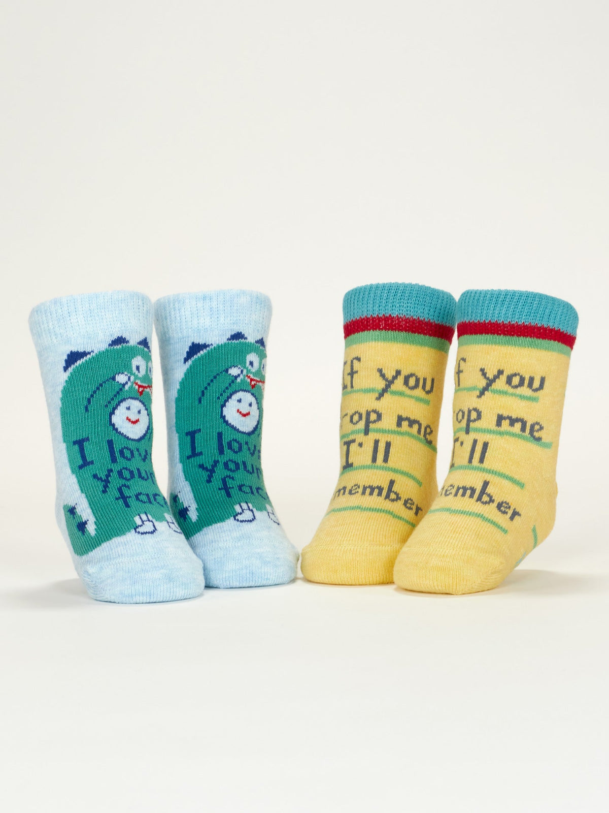 Two pairs of Blue Q's “I Love Your Face/If You Drop Me I'll Remember” baby socks are shown upright: one blue pair with a cartoon bear and message, the other yellow with green trim and a playful phrase. Both are organic cotton.