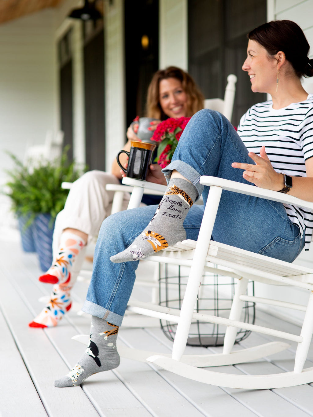 Two women sit on a cozy porch in white rocking chairs, wearing Blue Q's People I Love: Cats W-Crew Socks made of soft combed cotton and nylon spandex. They smile, chat, hold mugs, and are surrounded by green plants and a checkered pillow.