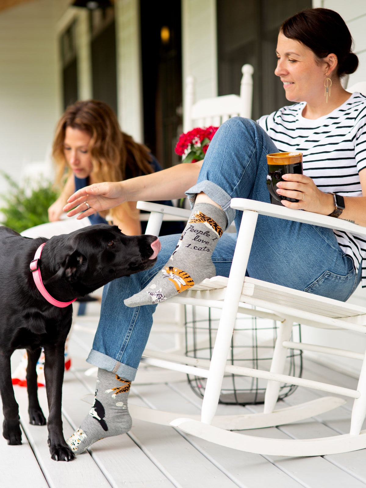 A woman in a striped shirt and jeans sits on a white rocking chair, wearing Blue Q's People I Love: Cats W-Crew Socks as she holds a drink and pets a black dog. Another woman sits nearby, both enjoying the cozy moment.