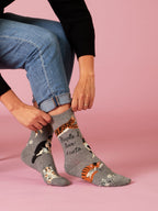 A person in blue jeans and a black sweater adjusts Blue Q’s People I Love: Cats W-Crew Socks—gray socks featuring cat illustrations and the words "People I love: 1. cats"—against a pink background.