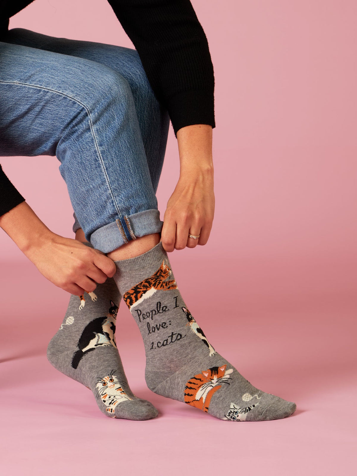 A person in blue jeans and a black sweater adjusts Blue Q’s People I Love: Cats W-Crew Socks—gray socks featuring cat illustrations and the words "People I love: 1. cats"—against a pink background.