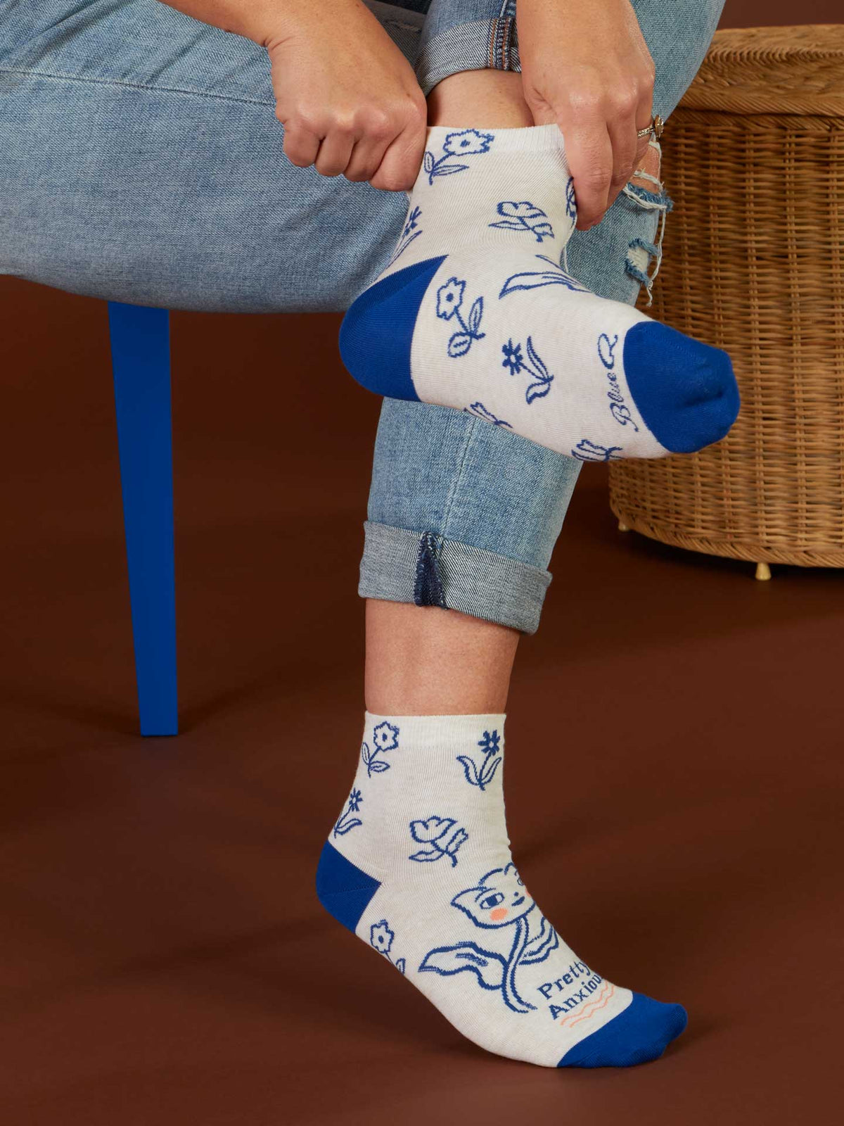 A person in Pretty Anxious W-Ankle Socks by Blue Q—soft combed cotton with blue toes, heels, and floral designs—sits on a blue chair rolling up their jeans. These cozy socks fit women's shoe size 5-10. A wicker basket is in the background.