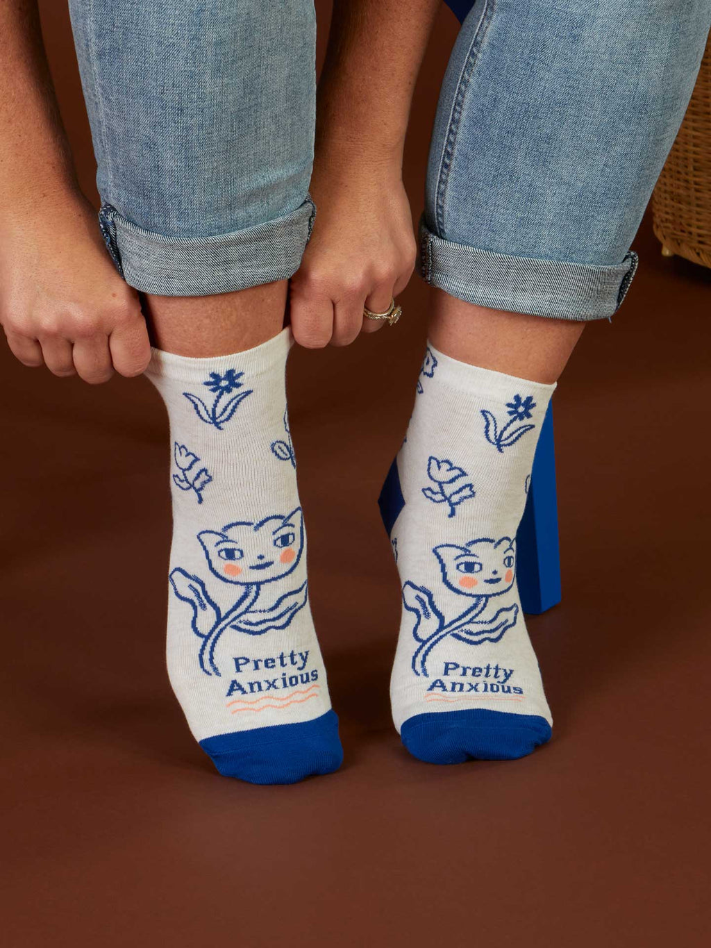 A person wearing Blue Q Pretty Anxious W-Ankle Socks—women's combed cotton socks with blue toes, cartoon cat, flowers, and "Pretty Anxious" text—sits holding their rolled-up jeans. Fits women's shoe size 5–10.