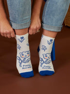 A person wearing Blue Q Pretty Anxious W-Ankle Socks—women's combed cotton socks with blue toes, cartoon cat, flowers, and "Pretty Anxious" text—sits holding their rolled-up jeans. Fits women's shoe size 5–10.